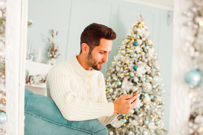 Young man using mobile phone against christmas tree