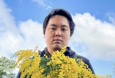 Portrait of young man holding golden wattle flowers against cloudy sky.