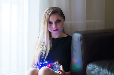 Portrait of mid adult woman holding illuminated lighting equipment while sitting by window at home