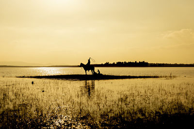 Silhouette person on shore by lake against sky during sunset