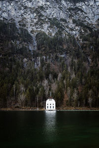 Picturesque scenery of lonely cabin located on shore of lake on background of coniferous forest and rocky mountains