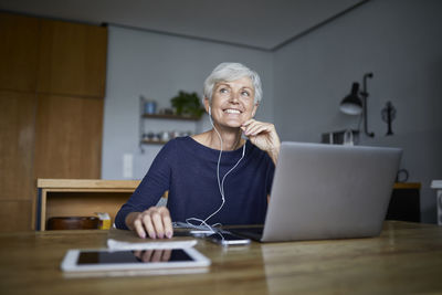 Active senior woman listening to music on smart phone and using laptop while looking away at home