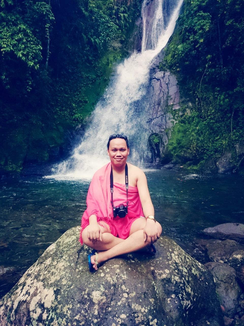 water, lifestyles, person, young adult, portrait, leisure activity, looking at camera, young women, casual clothing, rock - object, smiling, full length, front view, waterfall, standing, sitting, tree, happiness