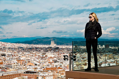 Young woman wearing warm clothing while standing on balcony against cityscape