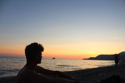 Young man looking away while sitting at beach against sky during sunset