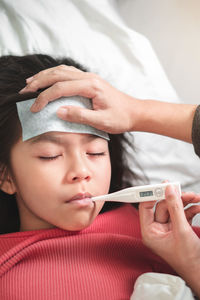 Cropped hand holding thermometer while girl resting on bed