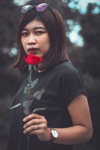 Portrait of young woman holding rose while standing against trees