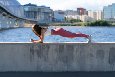 Full length of woman exercising by river against cityscape