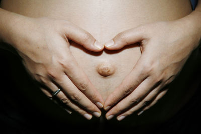 Midsection of pregnant woman making heart shape on abdomen against black background