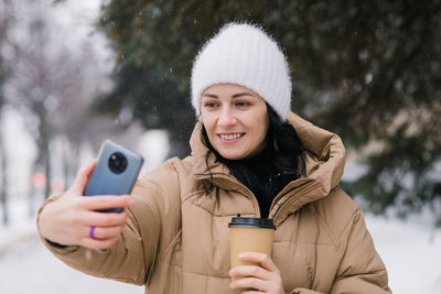 A cute girl laughs and takes a selfie outside in winter. hold a disposable cup of coffee