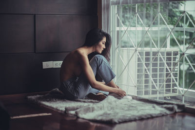 Shirtless young woman sitting by window at home