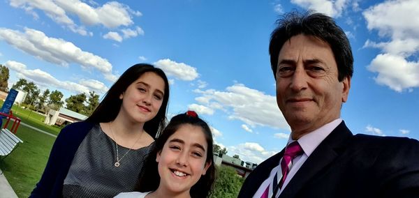 Portrait of grandfather standing with granddaughters at park against blue sky