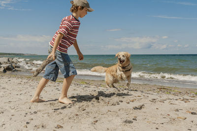 Boy playing with dog on sea shore at beach against sky