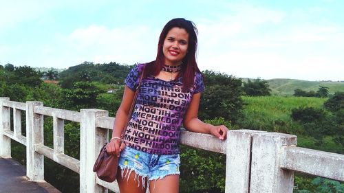 Portrait of smiling young woman standing by railing against sky