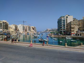 People on road by buildings against clear blue sky