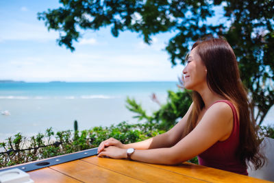 Young woman using phone on table by sea against sky