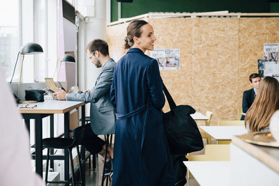 Smiling businesswoman with bag looking away while standing amidst colleagues in coworking space
