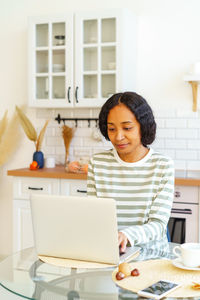 African-american female focused on remote work while sitting in kitchen and using laptop