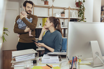 Smiling mother at desk looking at father holding baby in home office