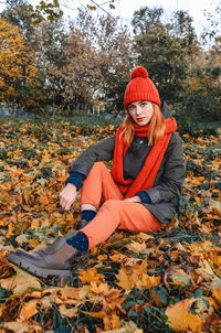 Autumn portrait of cute girl in orange warm knitted hat and orange scarf sitting in fallen leafs