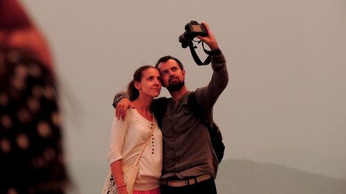 Friends taking selfie through camera while standing against sky during foggy weather