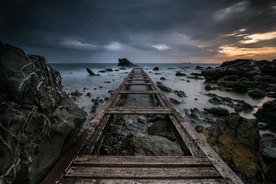 Damaged boardwalk at sea shore against cloudy sky during sunset