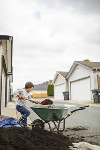 Young boy lifting shovel full of soil into wheelbarrow in back alley