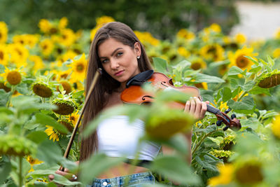 Young slim brunette with long hair standing in sunflower field and playing violin