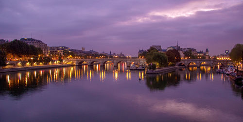 Illuminated bridge over river by buildings against sky at dusk