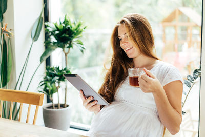 Charming pregnant woman drinks a drink and looks at the screen of a computer tablet.