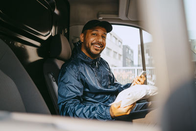 Portrait of smiling male delivery person holding parcel and smart phone while sitting in van