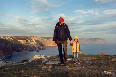 Man with a boy child walking on a mountain by the sea in autumn in a yellow vest about a hat