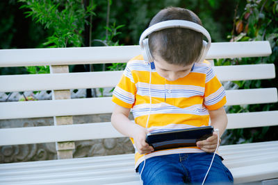 Cute boy listening music while using mobile phone on bench