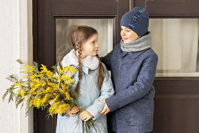 A couple of a boy and a girl of schoolchildren in spring coats with a bouquet of mimosa in  hands 