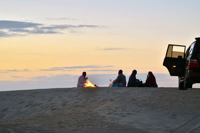 People sitting on beach by sea against sky during sunset