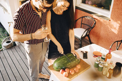 High angle view of man with wineglass looking at female friend cutting watermelon in party