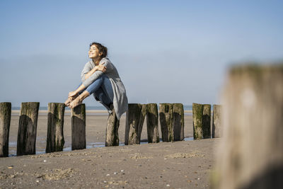 Smiling young woman sitting on wooden posts at beach against sky