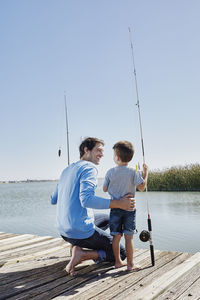 Smiling father looking at son standing with fishing rod on pier