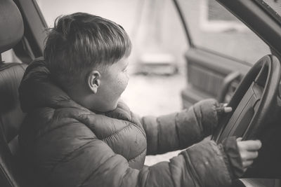 Side view of boy wearing hooded jacket while sitting on driver seat in car