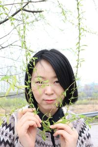 Portrait of smiling young woman holding sunglasses against trees