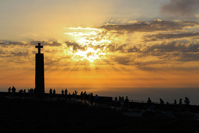 Silhouette people around cross on hill by sea against cloudy sky during sunset