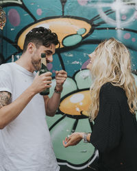 Man holding beer bottle by woman while standing against wall