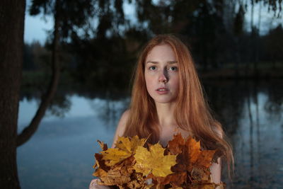 Portrait of young female model covered with autumn leaves against lake at dusk