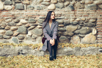 Full length of young woman sitting against wall during autumn