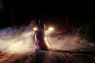 Full length of woman standing against car on dirt road at night