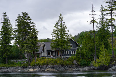 House by river amidst trees and buildings against sky