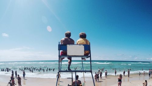 Rear view of man and woman sitting on lifeguard chair at beach against sky