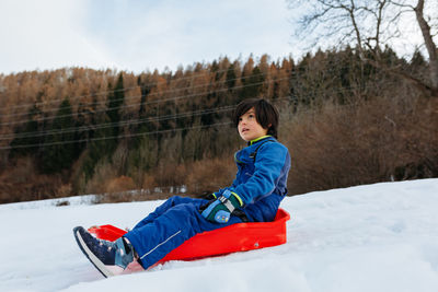 Child in blue winter clothes sliding down the snowy hill in red sled