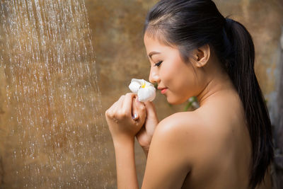 Rear view of topless young woman holding flower while taking bath in shower