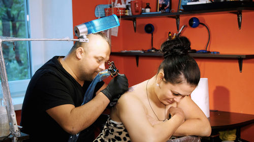 In a tattoo salon , a specialist is doing a tattoo on woman's back, a floral ornament. a man works